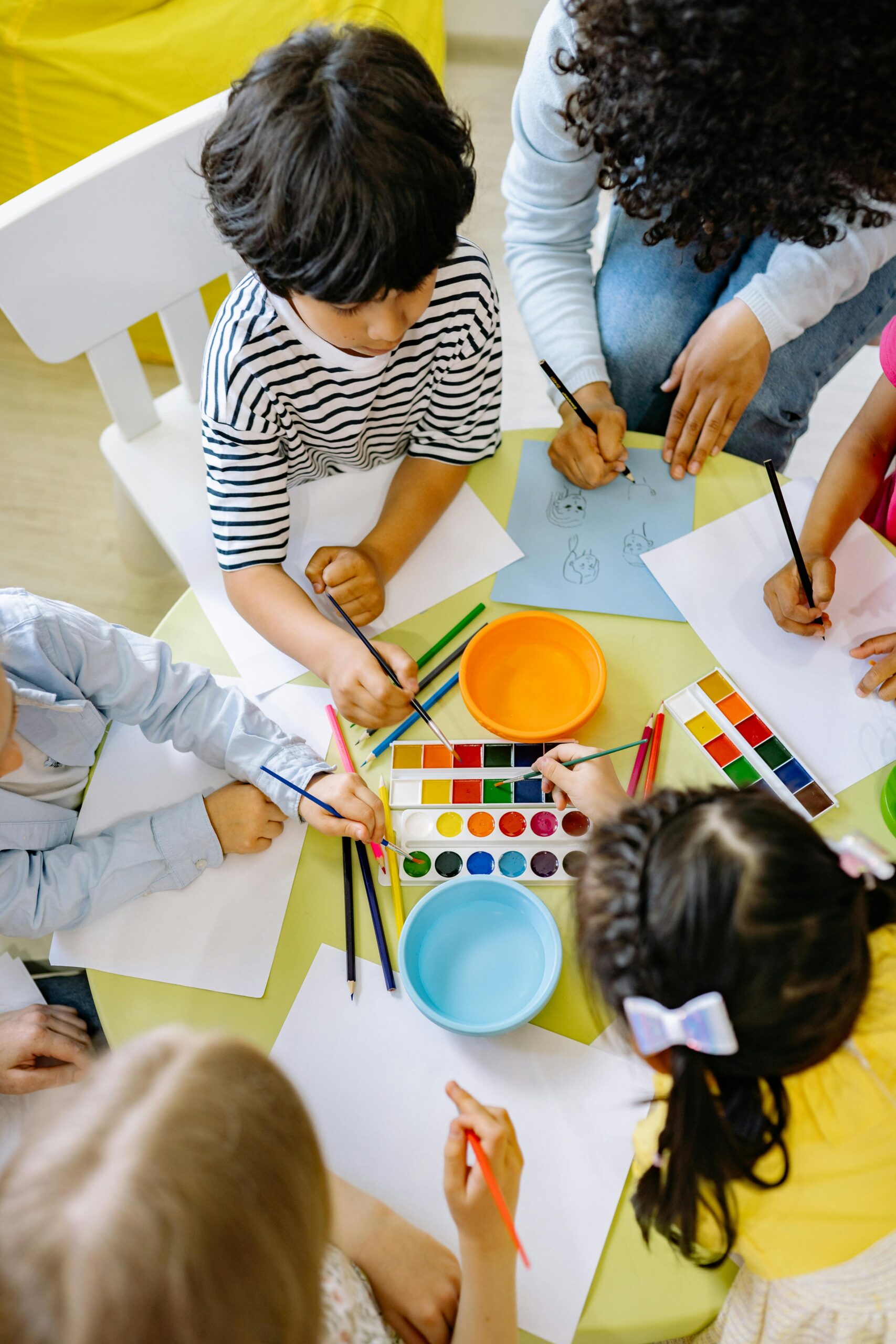 Children and an adult painting and drawing at a yellow table with art supplies in the center.