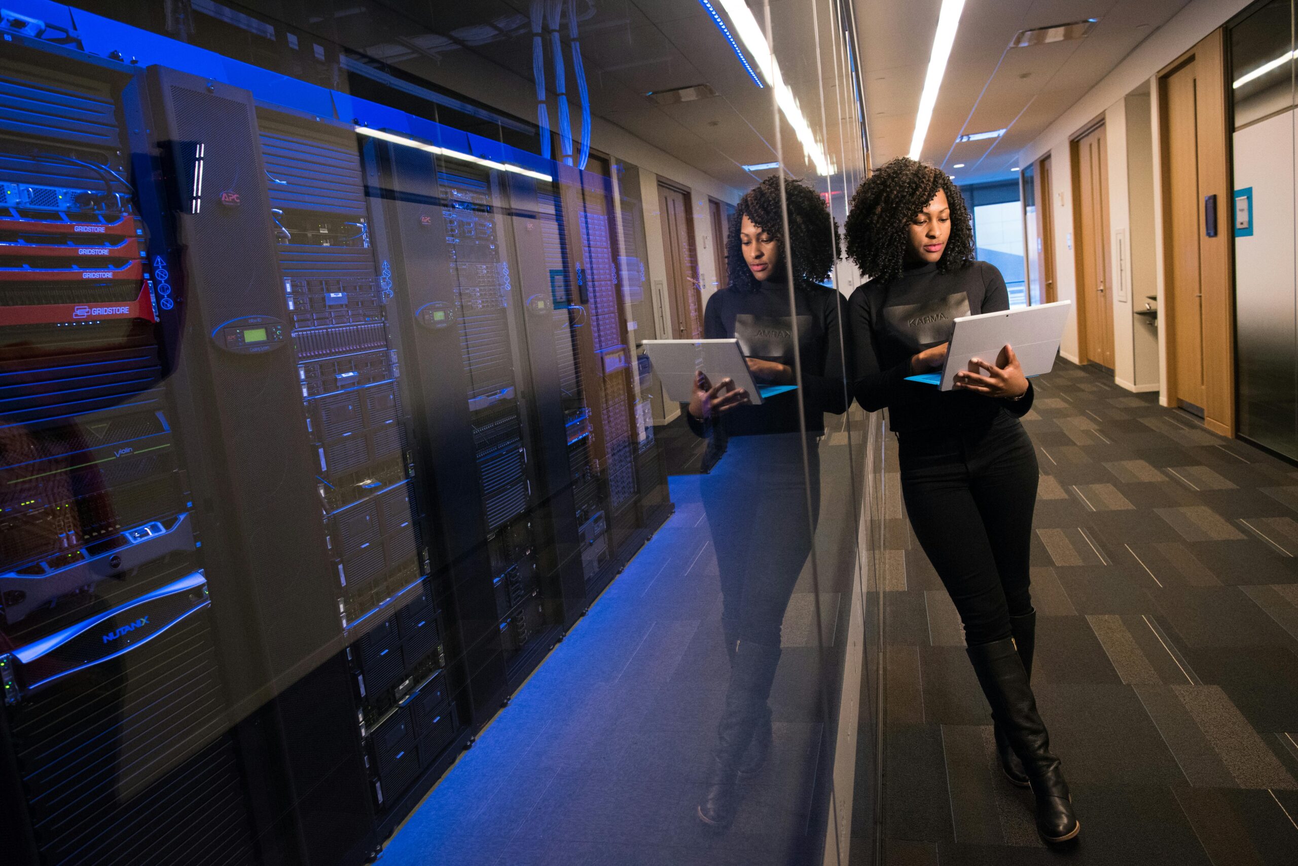 Woman in hallway, looking at her laptop, leaning against server room glass.