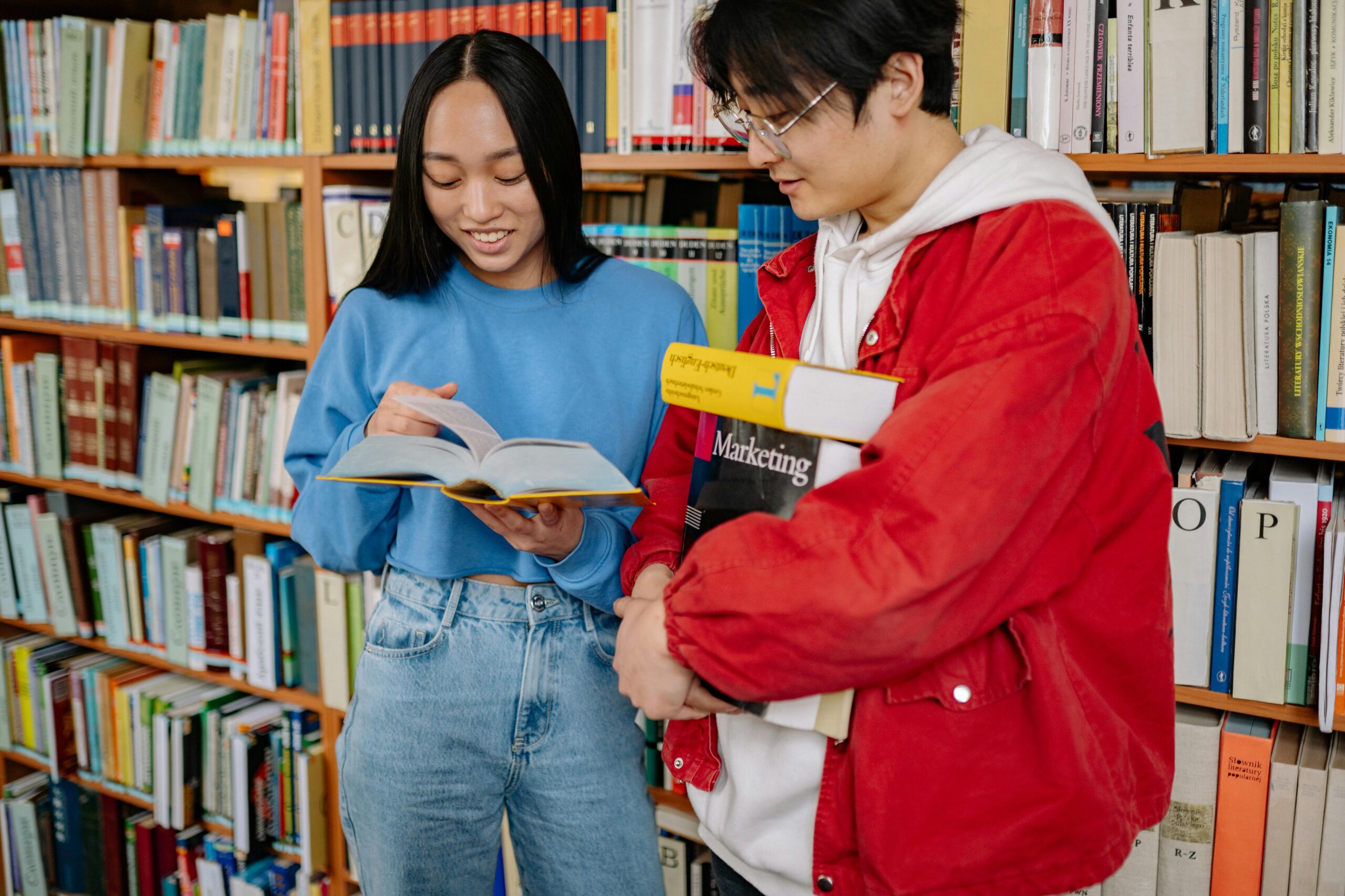 Woman flipping through a book while man with a stack of books looks on, in front of a bookshelf.