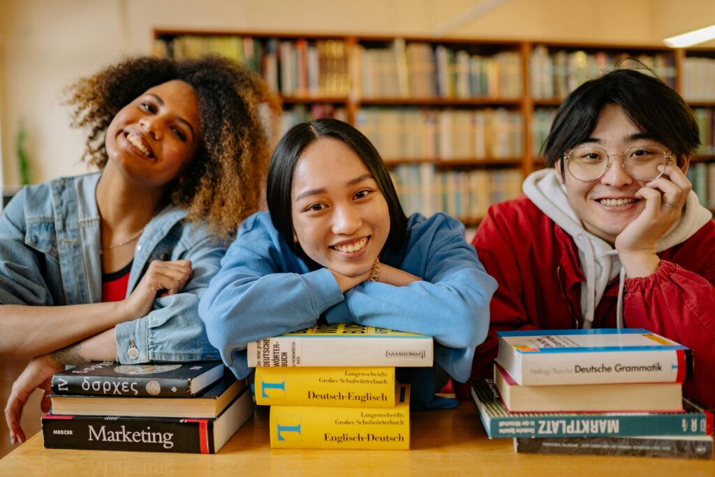 A group of three smiling young adults sitting at a library table. There are stacks of books in front of them. There are shelves behind them, filled with books. 