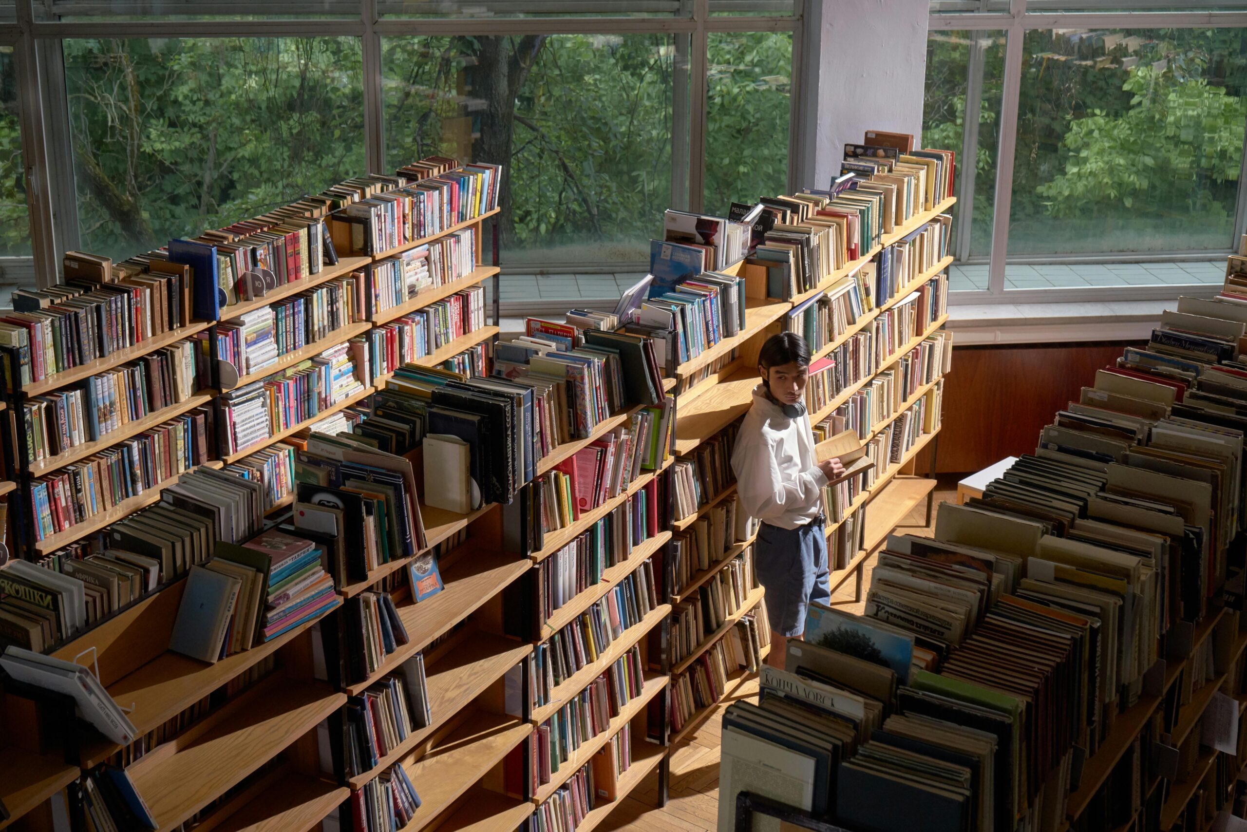 View of library rows with books, windows showing trees, and a person holding a book.