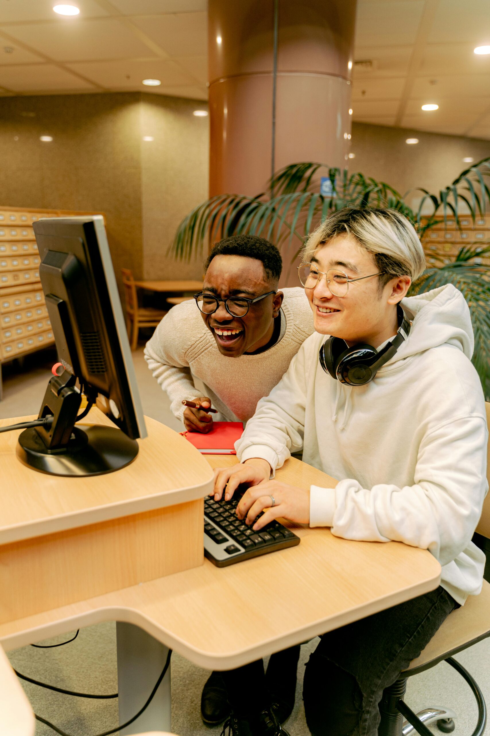 Two smiling men sitting in a library in front of a computer. 