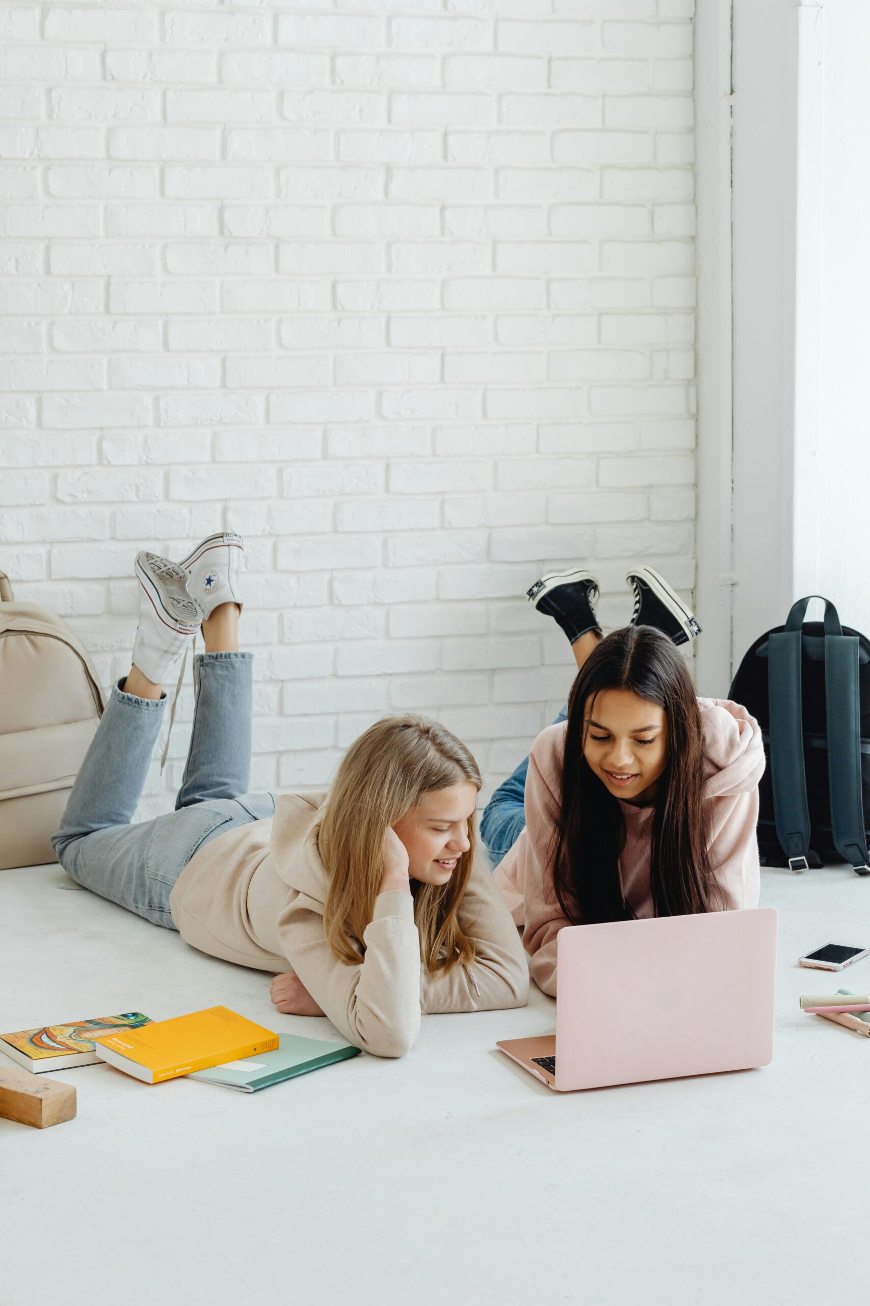 Two smiling teenage girls on the floor with a pink laptop, surrounded by bookbags and notebooks.